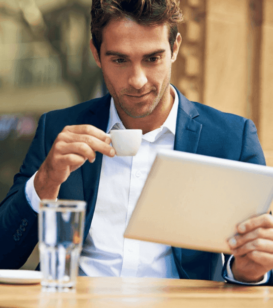 Professional man sips coffee while reading a tablet at an outdoor cafe.