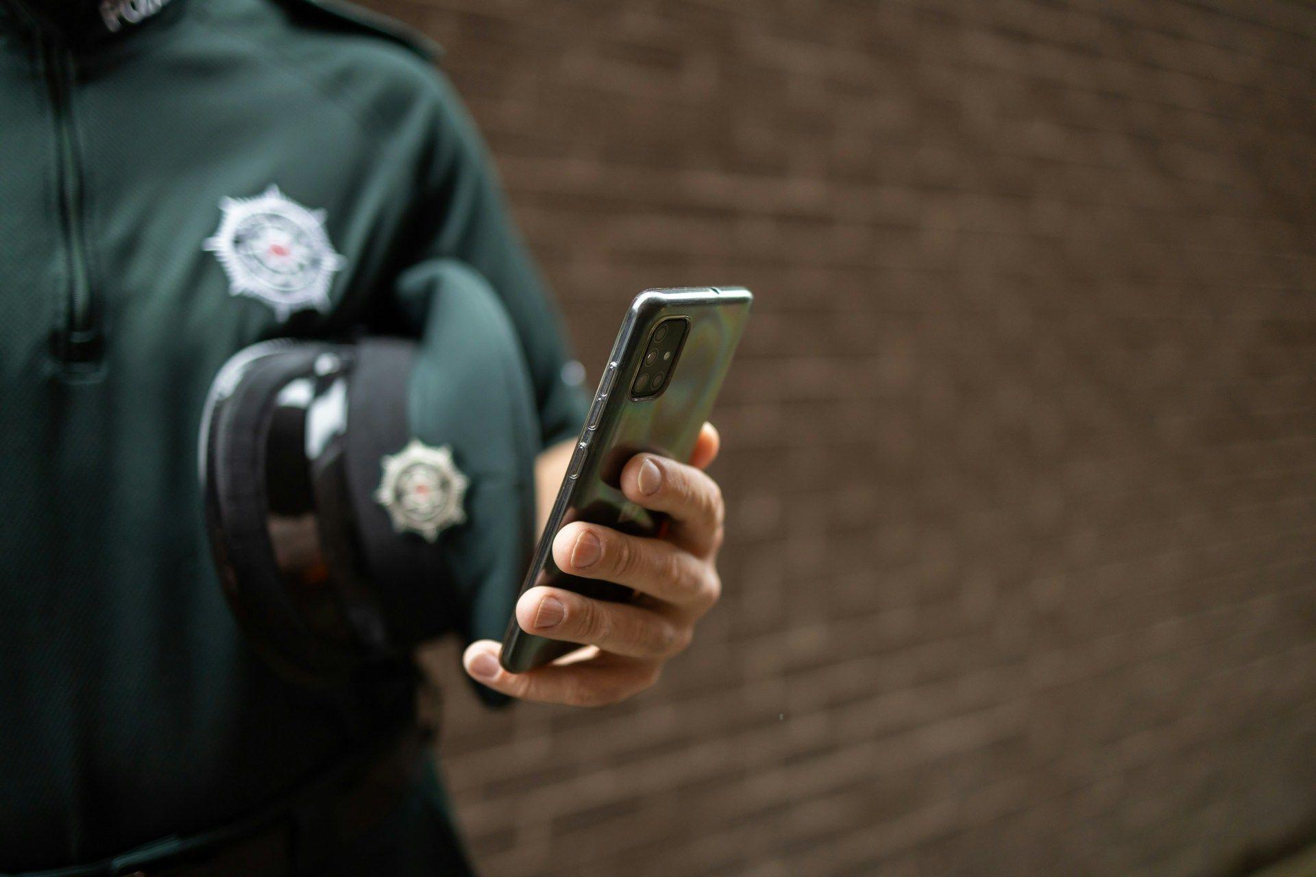 A person in uniform holds a smartphone and a peaked cap.