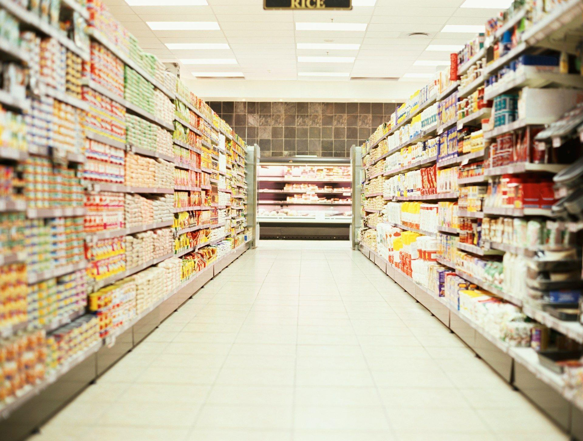 Supermarket aisle with stocked shelves of various products stretching into the distance.