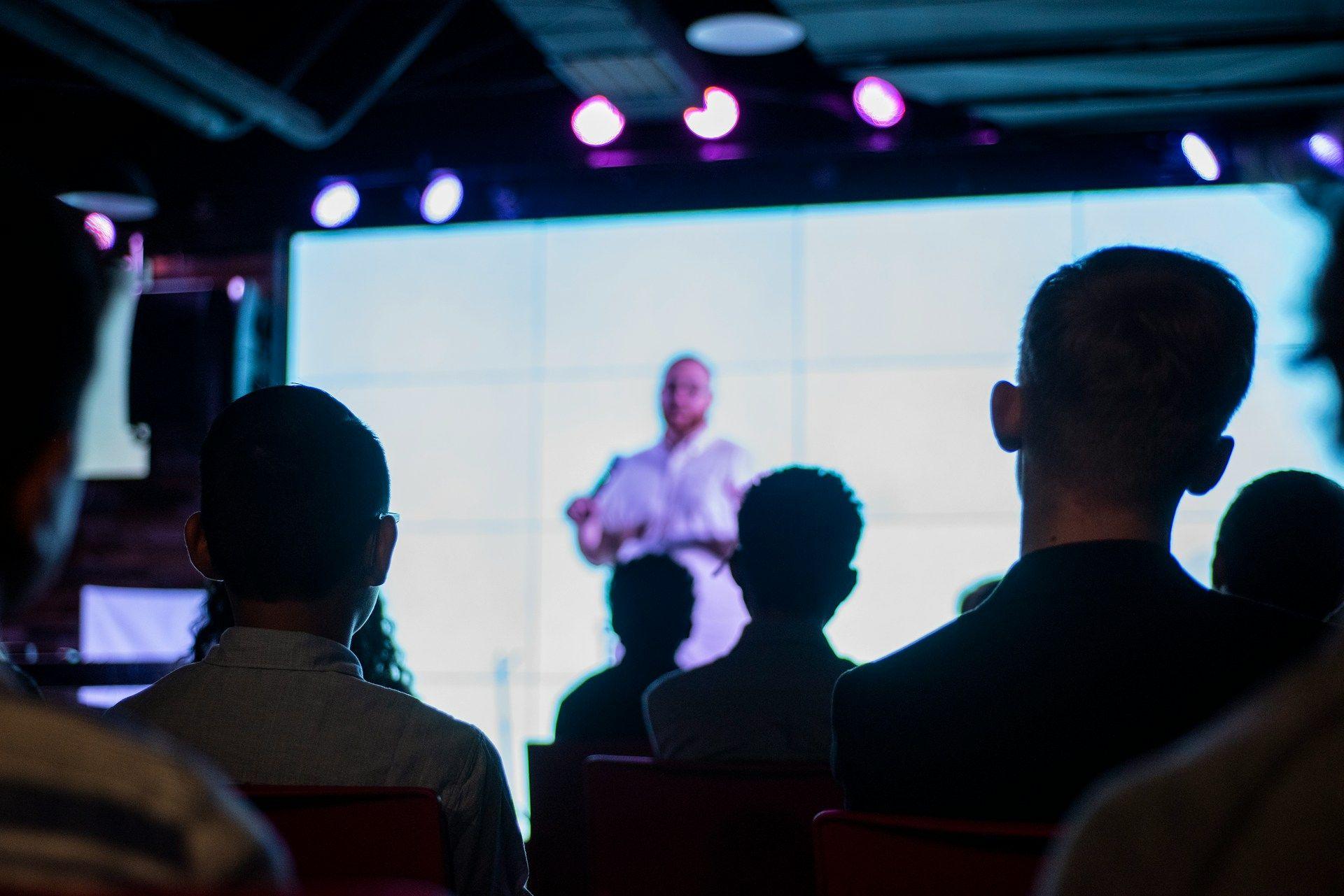 Audience watching a speaker present on a brightly lit stage screen.